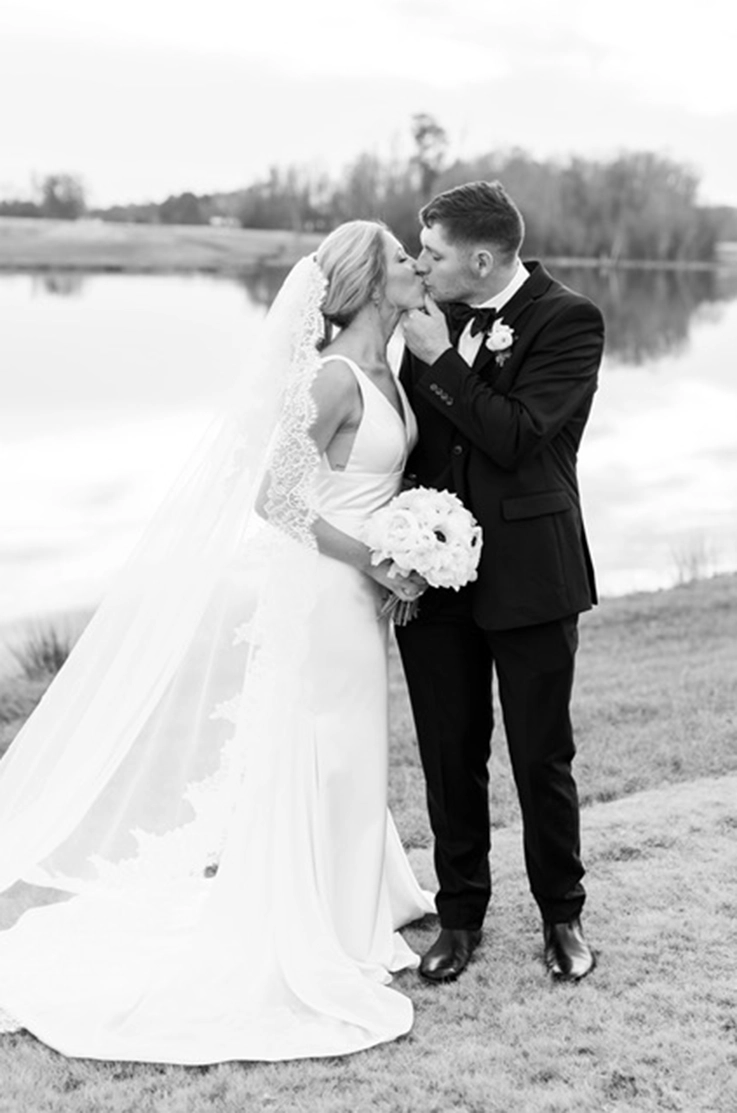 Groom kisses bride by a lakeside in a sleeveless satin gown with an empire waist and lace-edged veil, in black and white.
