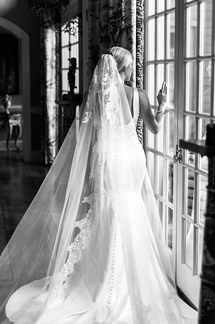 Bride looks out French doors in a sleeveless fit and flare gown with buttons down to the train and a lace-trim veil. 