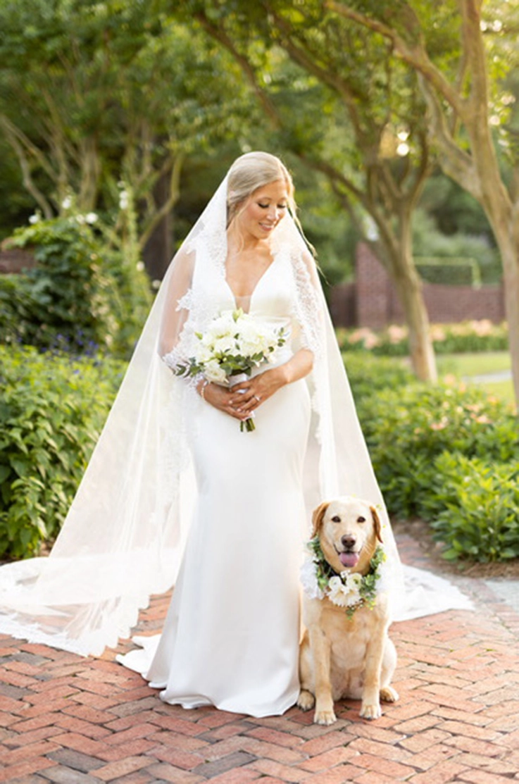  Bride poses in a satin fit and flare wedding dress with a plunging neck and lace-trim veil, next to her dog on a brick path.