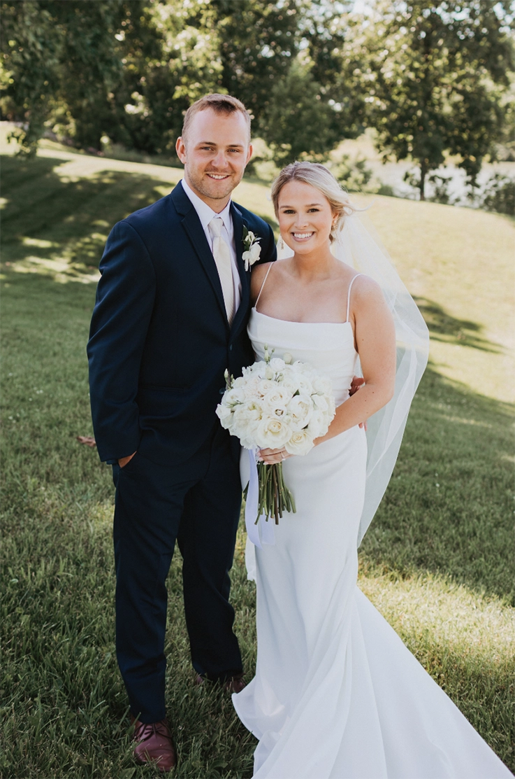  Bride in a crepe fit and flare gown with a scoop neck, pleating and a veil, holds a white bouquet and smiles next to groom.