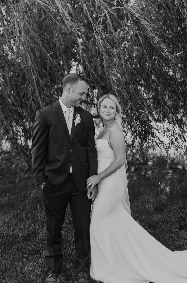 Bride in a scoop neck crepe gown with a pleated bodice and a veil, poses with groom under willow trees in black and white.