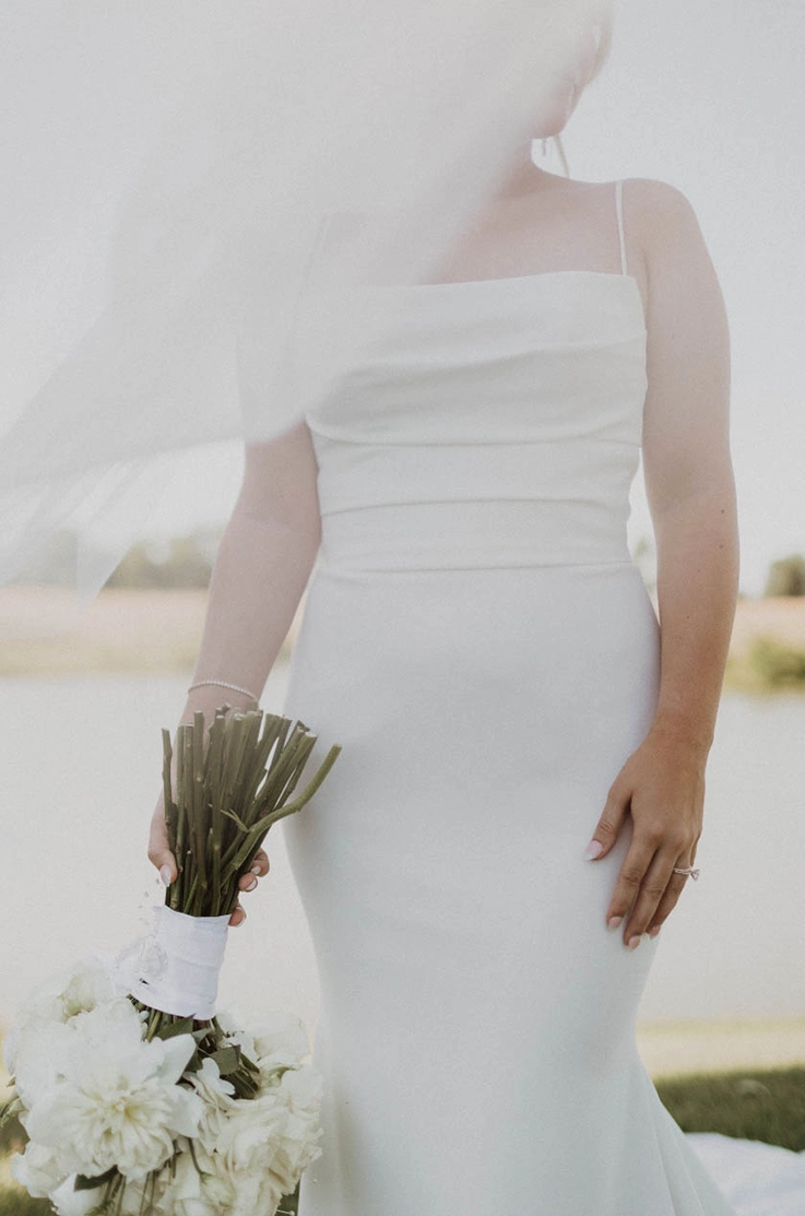 Close-up of bride in a crepe fit and flare wedding dress with pleating and a flowing veil, is holding a bouquet to the side. 