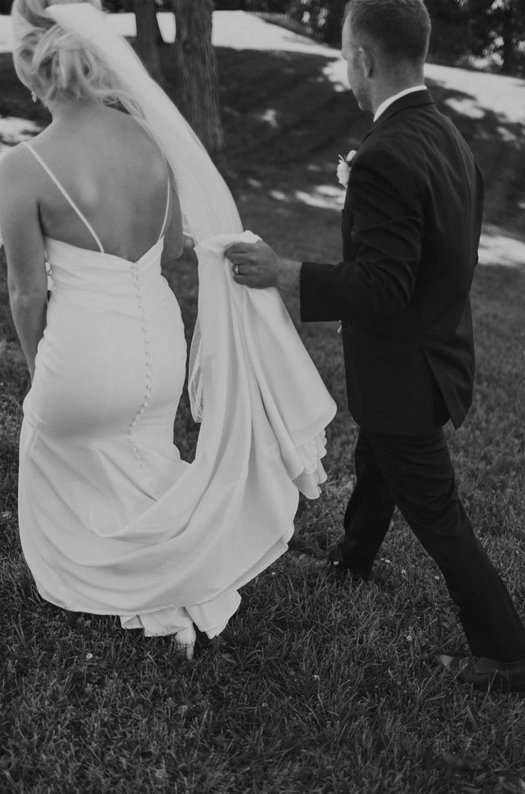Groom picks up bride's train in a low back crepe gown with a veil, low back and thin straps as they walk together on a lawn.