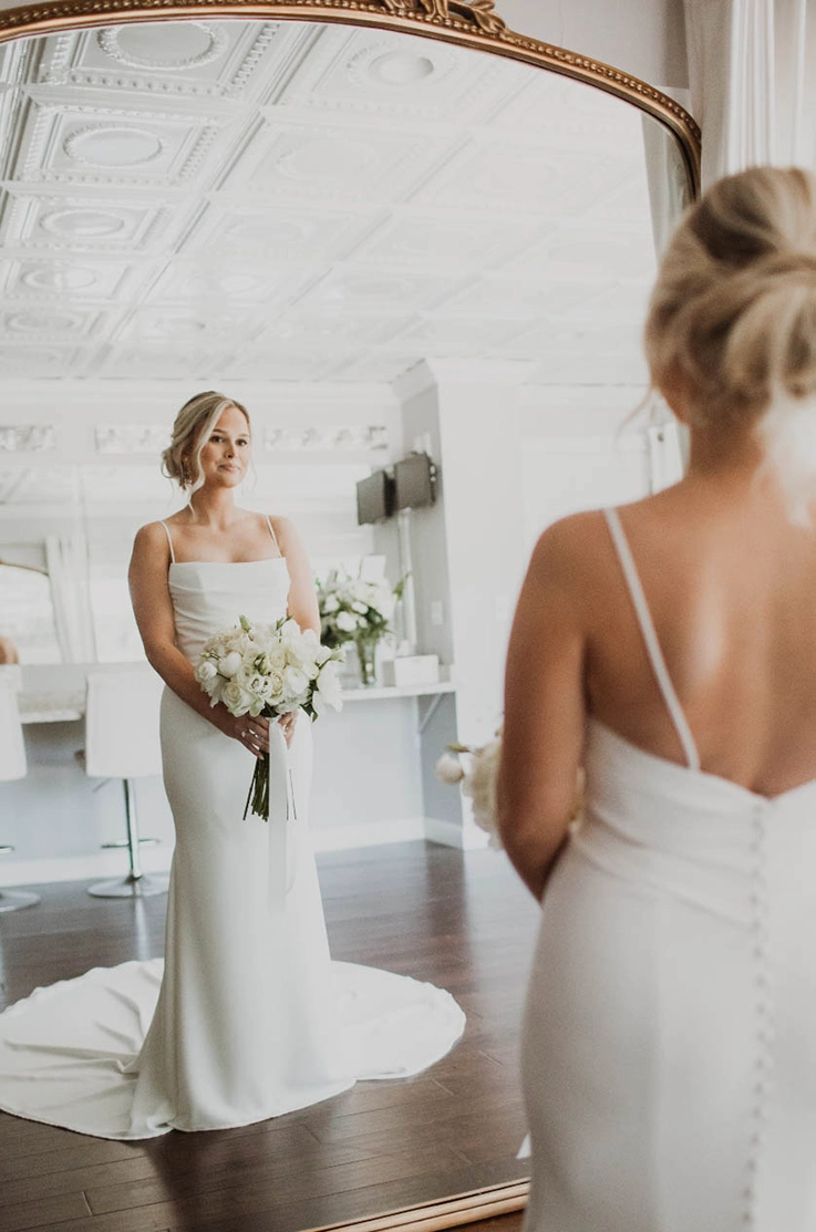  Bride in a crepe pleated bodice gown with a scoop neck and fit and flare skirt, looks at her reflection in a bridal suite mirror.
