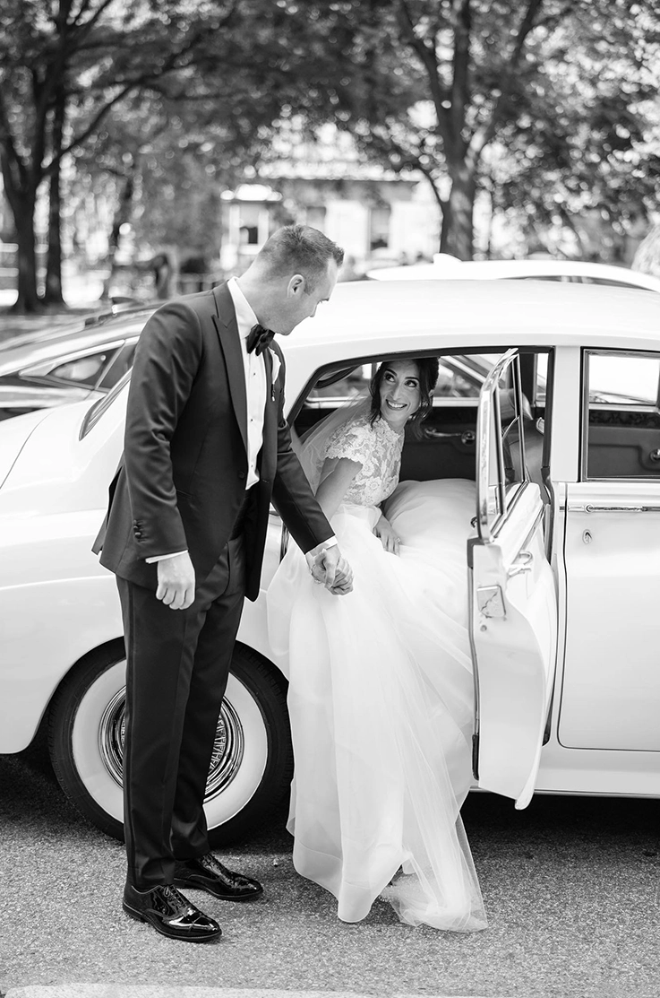 Bride in a lace and organdy gown with a jewel neckline and cap sleeves, exits a vintage car and looks up at her groom.
