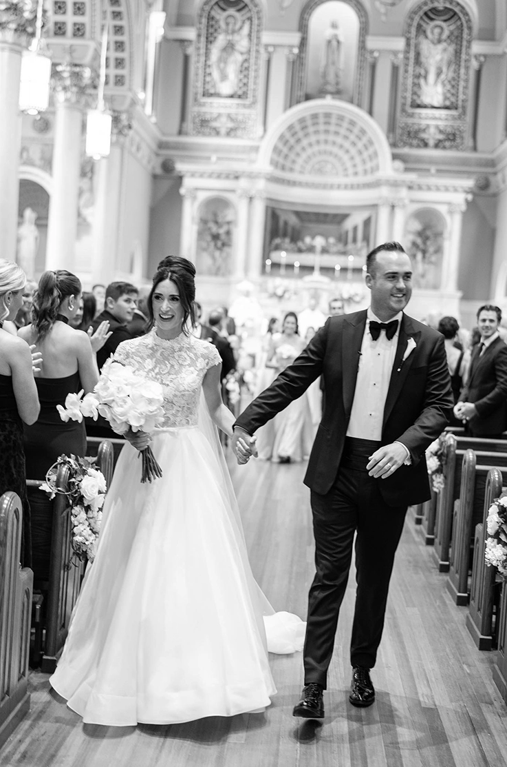 Bride in a lace bodice cap sleeve wedding dress with a full organdy skirt, walks down the aisle with groom in black and white.