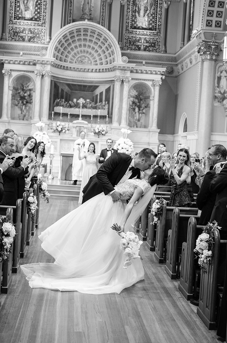 Groom dips his bride in a lace bodice gown with a veil and full organdy skirt, holding a bouquet in a grand cathedral.