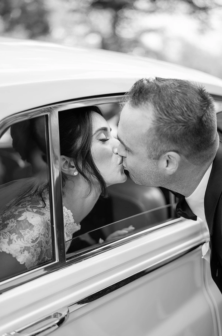 Bride in a lace jewel neck gown kisses gown in the window of a vintage car, and is shown in black and white.