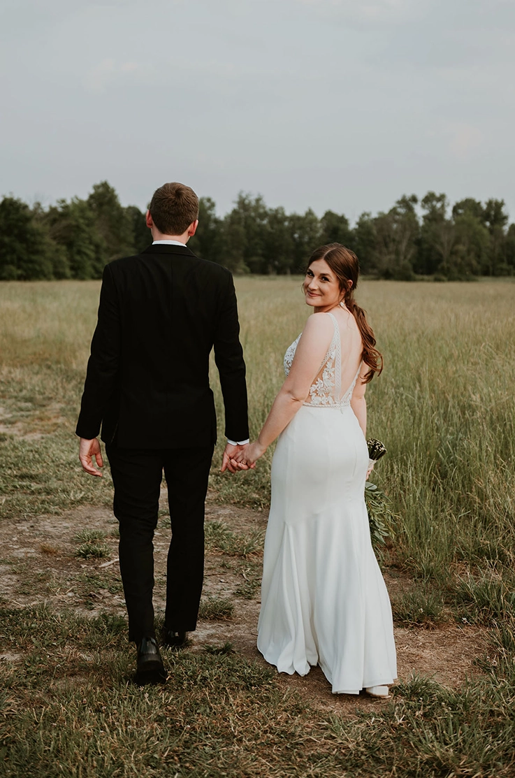 Bride looks past her shoulder in a deep-V back lace gown with a crepe skirt and walks hand in hand with the groom in a field.