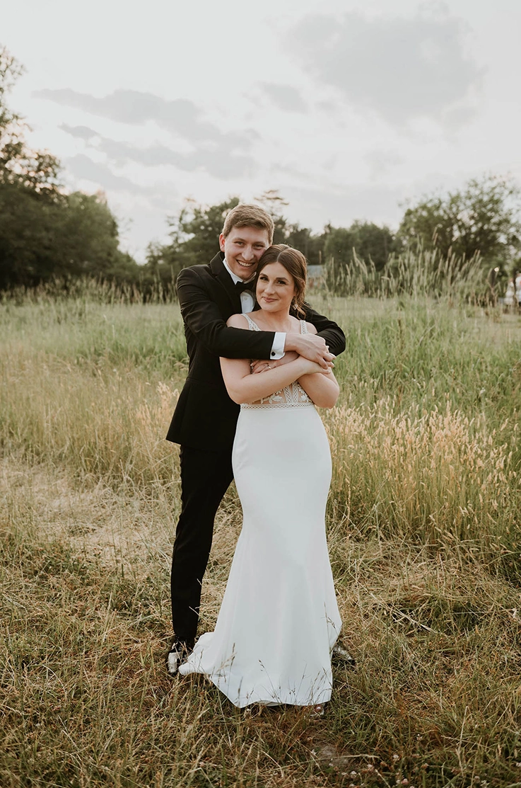 Groom holds his bride in an embrace in V-neck lace gown with a crepe fit and flare skirt in an open field.