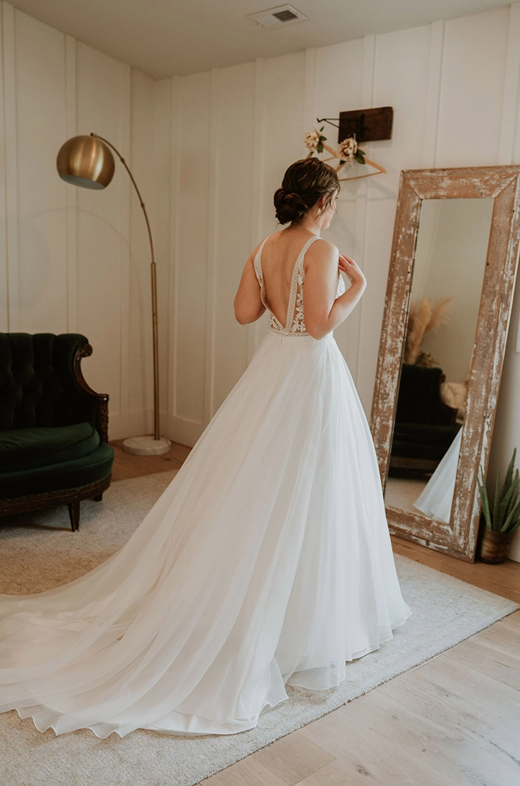 Bride in a lace bodice gown with a V-shaped back and flowing skirt, looks at her reflection in a mirror in a bridal suite.