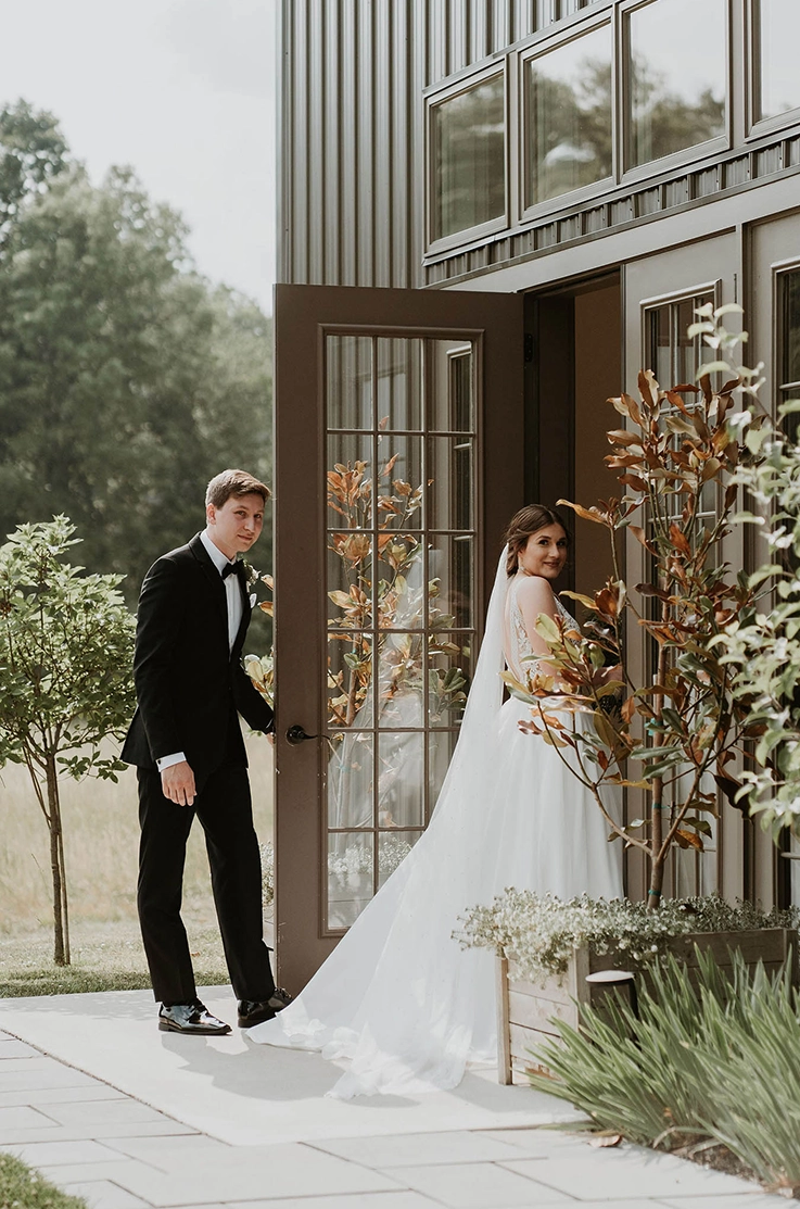 Bride in a sleeveless lace bodice wedding dress with a crepe skirt and veil, enters venue alongside her groom.