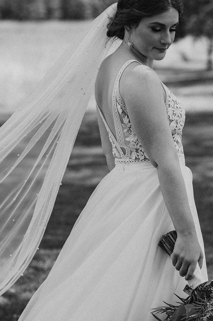 Close-up of bride posing in a lace and crepe gown with a pearl veil, and holds a bouquet to the side in black and white. 