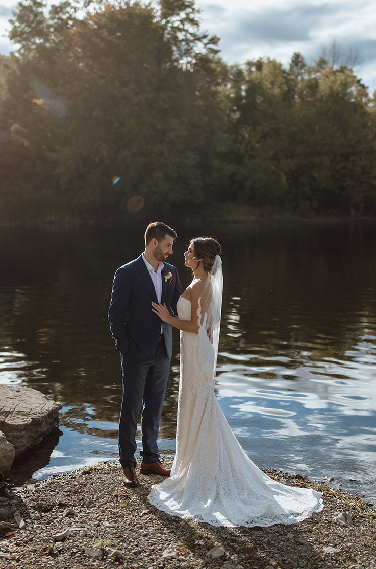 Bride in a strapless lace wedding dress with a tulle fit and flare skirt and veil, poses with groom on a rocky lakeshore.