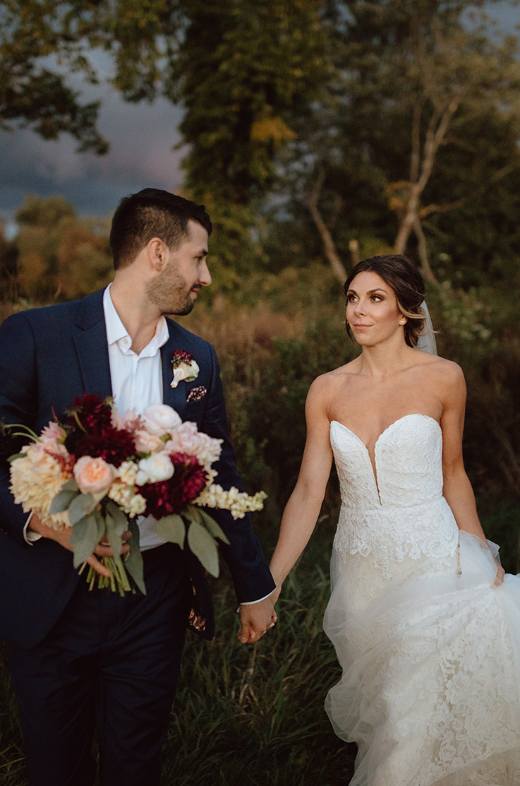Bride in a strapless lace and tulle gown with a sweetheart plunging neckline, walks hand in hand with her groom in a field.