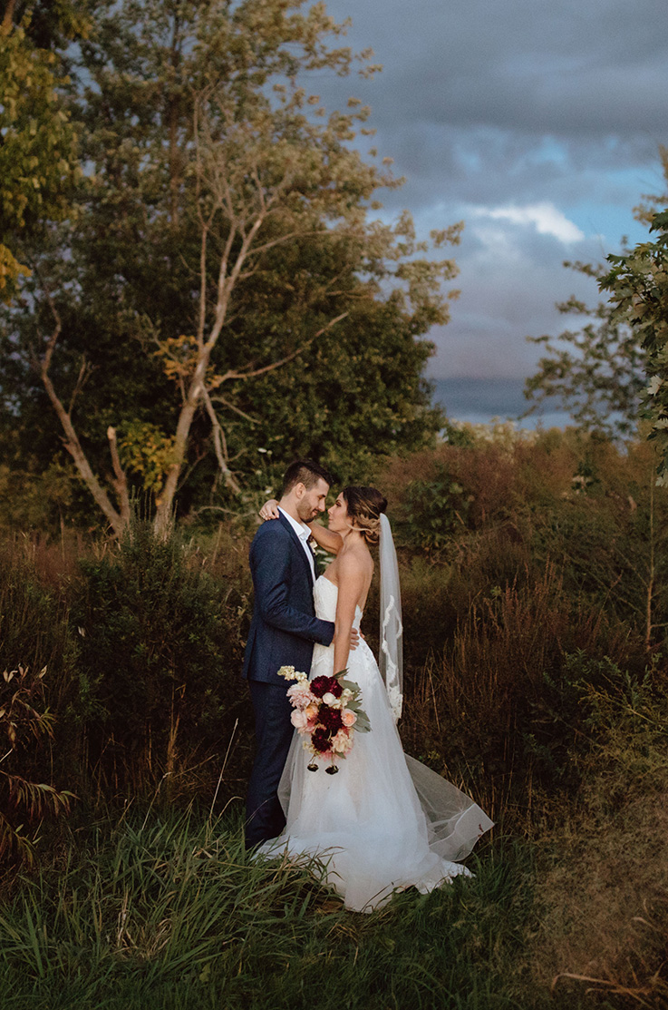 Grooms holds bride in an embrace in a strapless lace gown with a tulle overskirt and a veil, and holds a bouquet in tall grass.