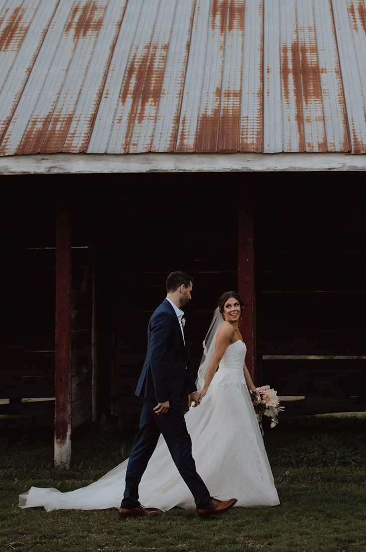 Bride glances back at groom past a rustic barn, wearing a strapless lace gown with a tulle skirt and a veil, holding a bouquet. 