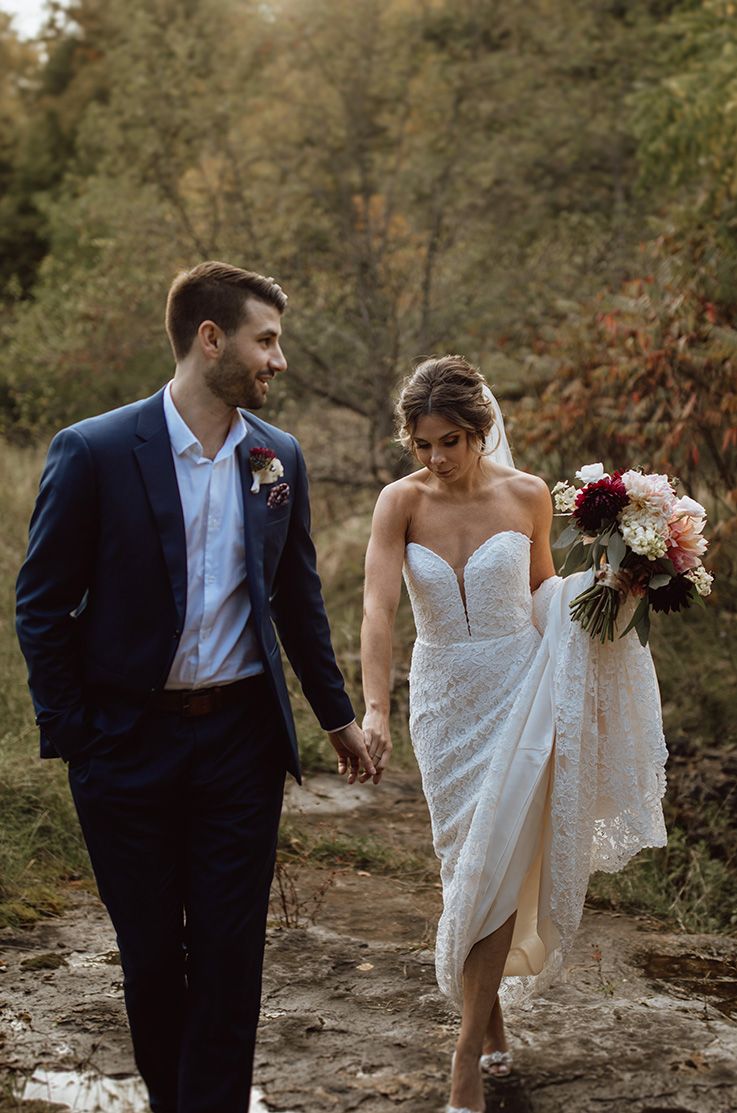 Bride picks up her train in a strapless lace gown with a sweetheart neck, and is walking hand in hand with her groom outdoors. 