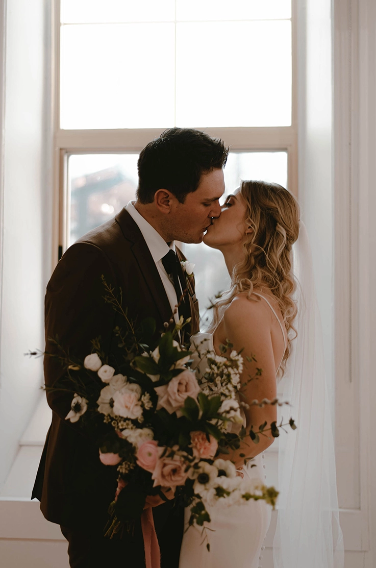 Groom kisses his bride in a fit and flare crepe gown with a veil and is holding a large bouquet by a bright window.