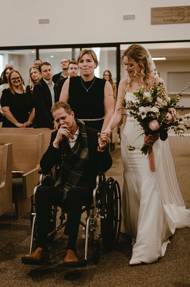 Bride in a crepe pleated bodice gown with a veil, holds a bouquet and walks down the aisle with her dad in a wheelchair.
