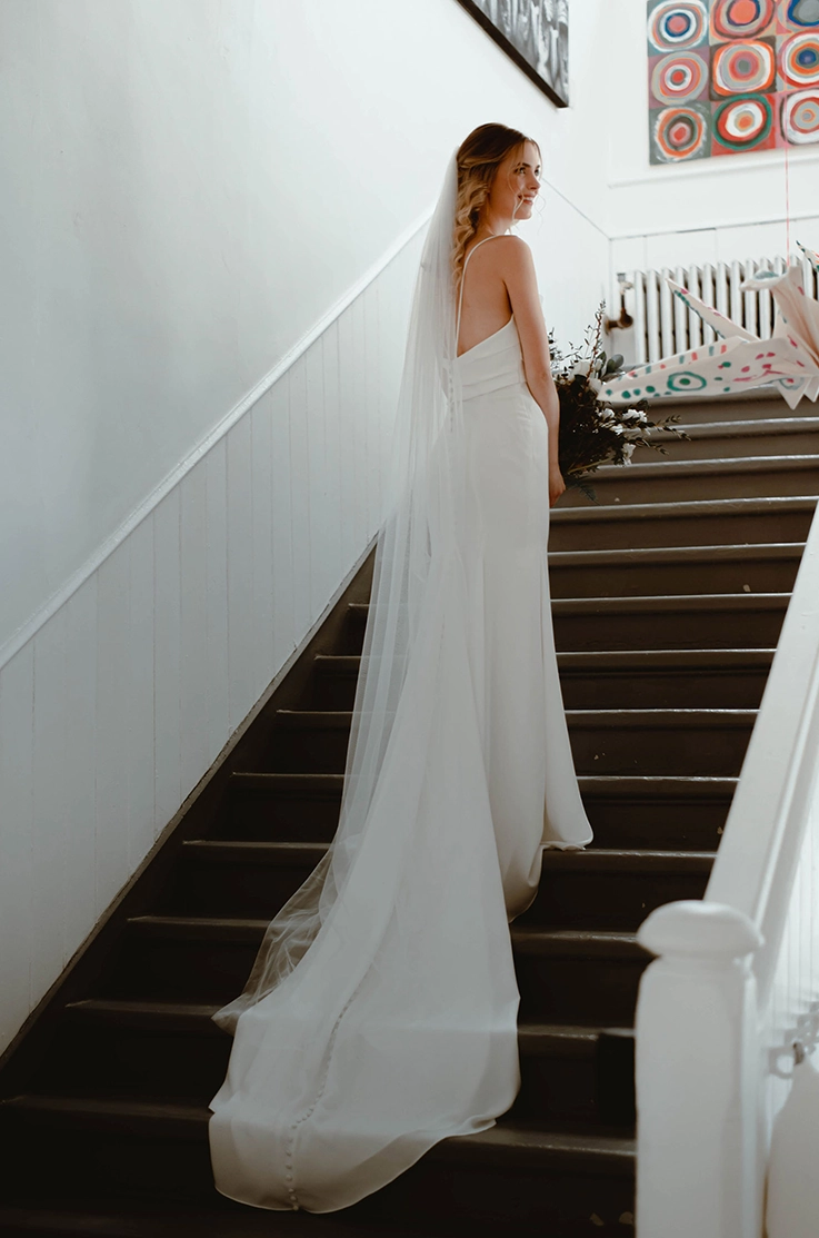 Bride in a crepe fit and flare wedding dress with a long veil and train with button detail, walks up a staircase indoors.