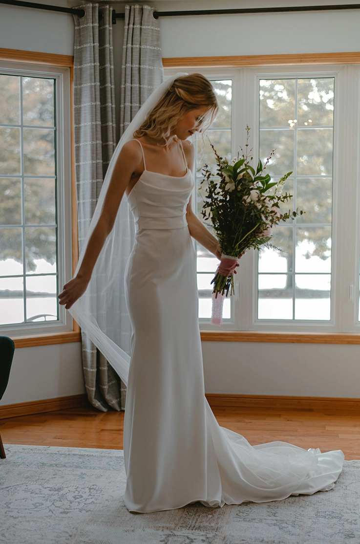 Bride in a fitted crepe gown with a draped scoop neckline and thin straps, picks up her veil and holds a bouquet near windows. 