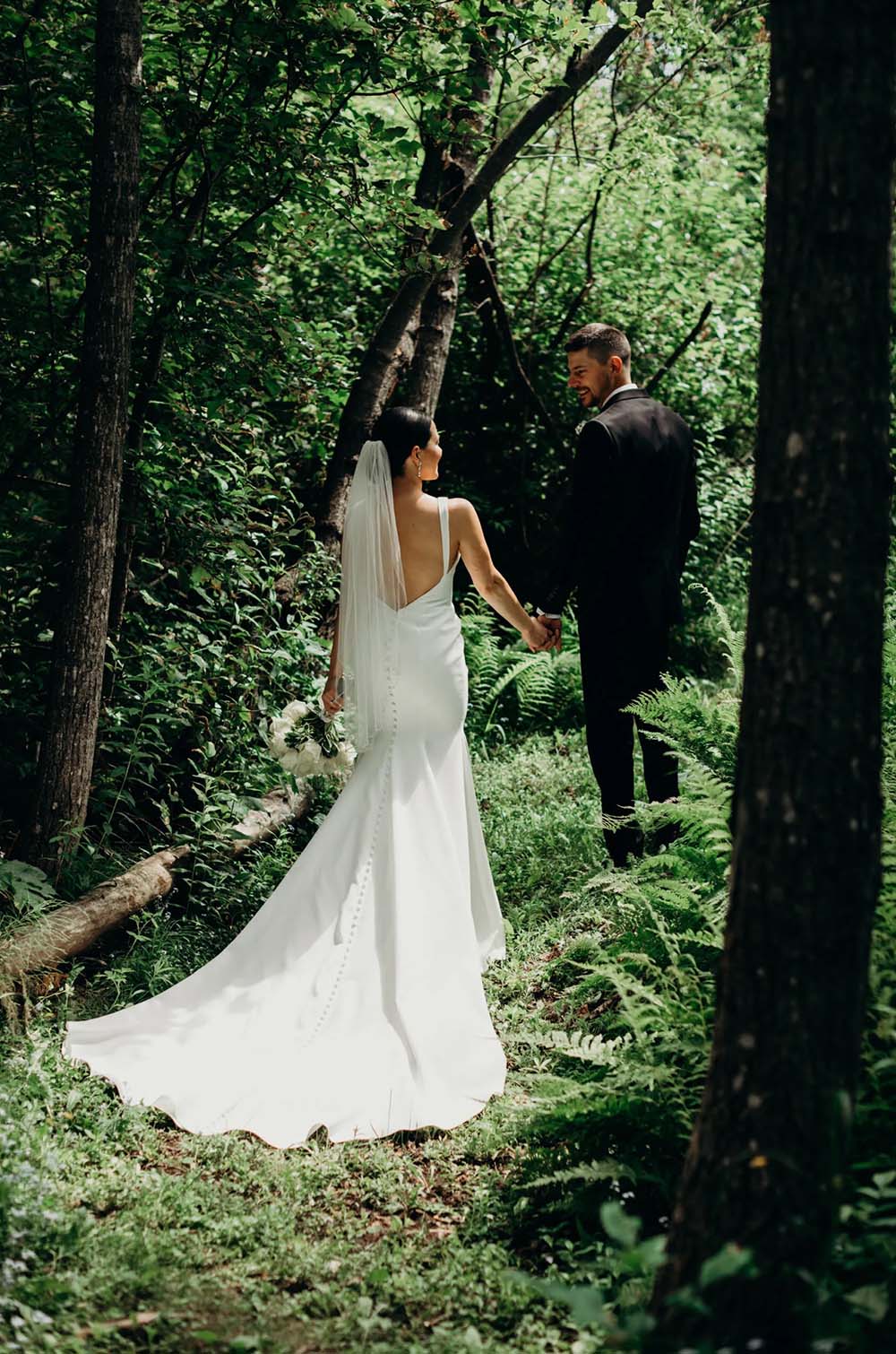 Groom holds bride's hand in a crepe deep-V back gown with buttons, and a veil, as they walk through a forest.
