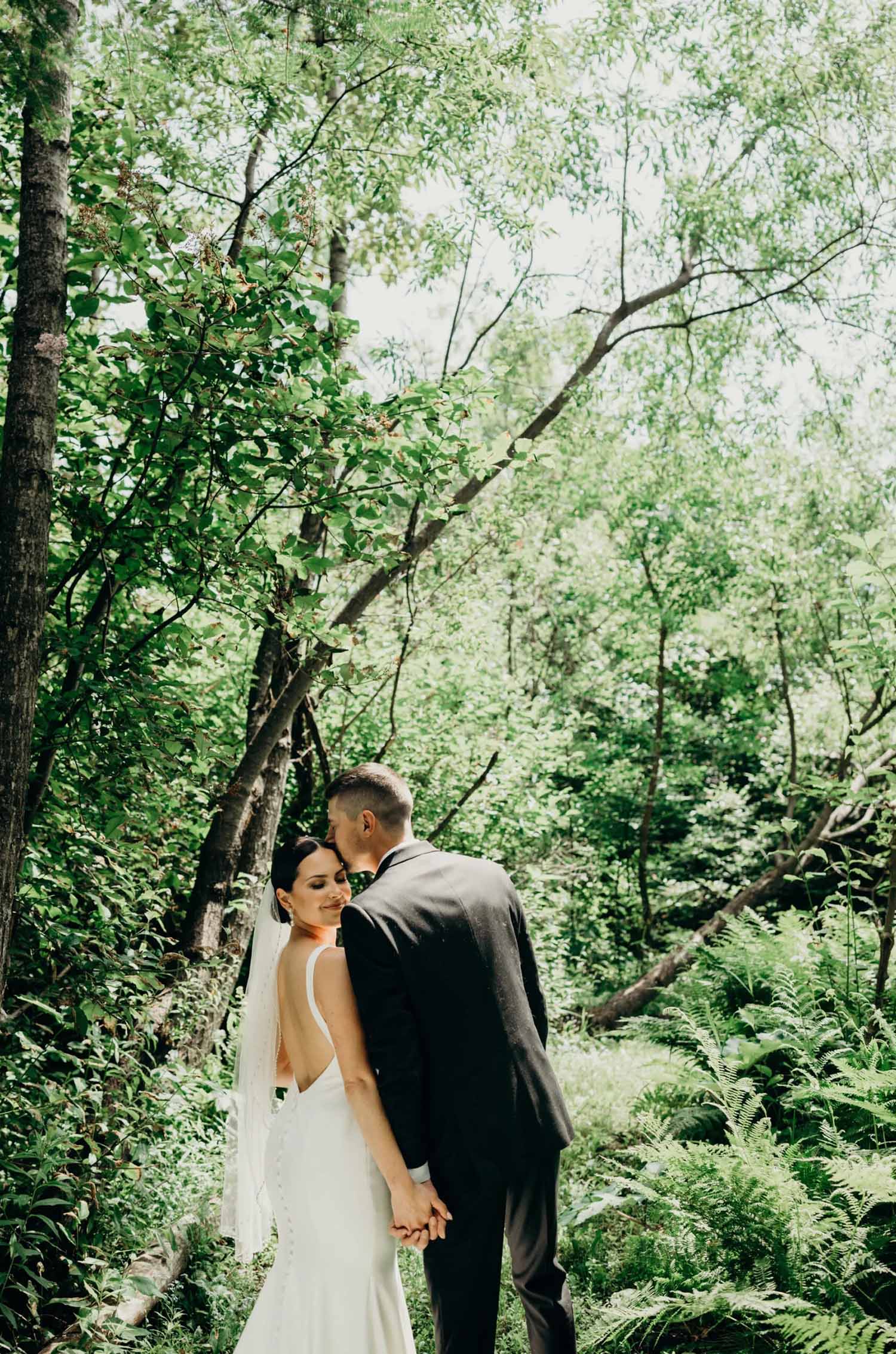 Groom kisses bride in a crepe fit and flare gown with a low back, buttons and a veil in front of trees.