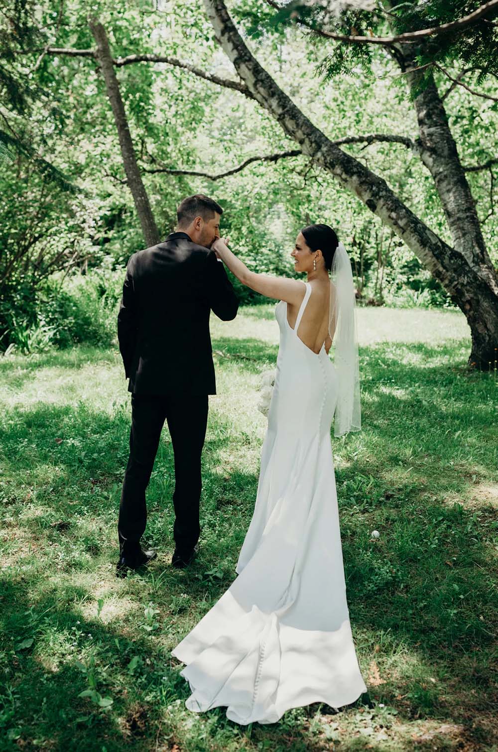 Bride in a fit and flare crepe wedding dress with a low buttoned back and a veil, poses with groom in a sunny forest area.