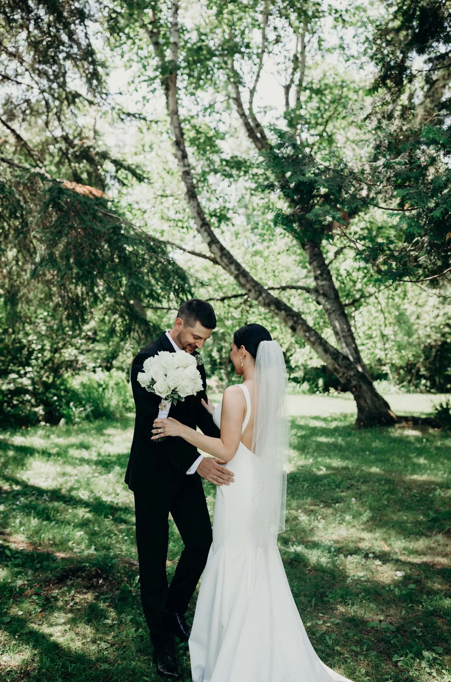 Bride in a fit and flare crepe gown with a low back and a veil, poses with her groom in a sunny forest clearing.
