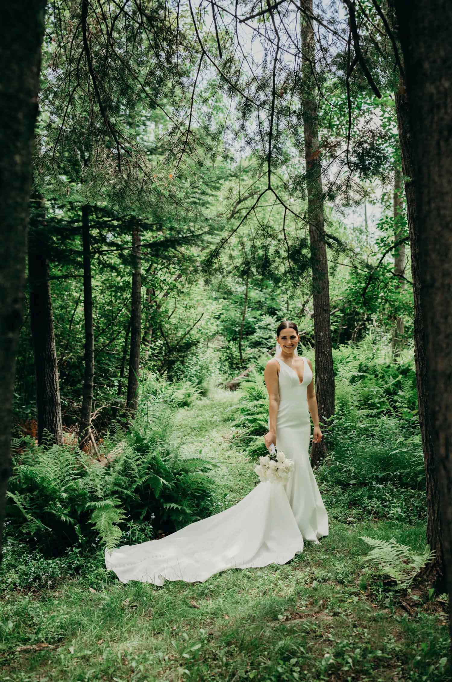Bride poses in a sleeveless crepe gown with a fit and flare skirt, veil and plunging neck, holding a bouquet on a forest path.