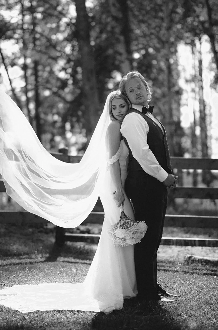 Bride in a crepe bodice gown with lace sleeves and a flowing veil, holds a bouquet and poses with groom in black and white.