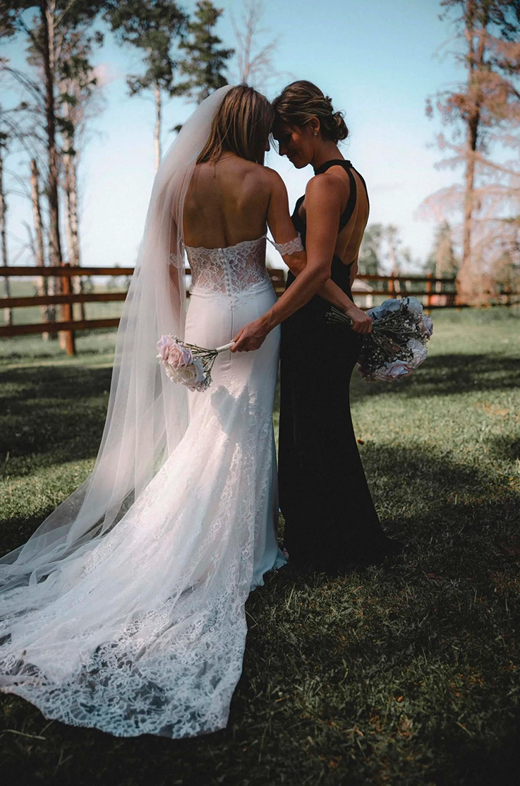 Bride in a lace corset back gown with sleeves and fit and flare crepe skirt with a veil, poses with a bridesmaid, in a field.