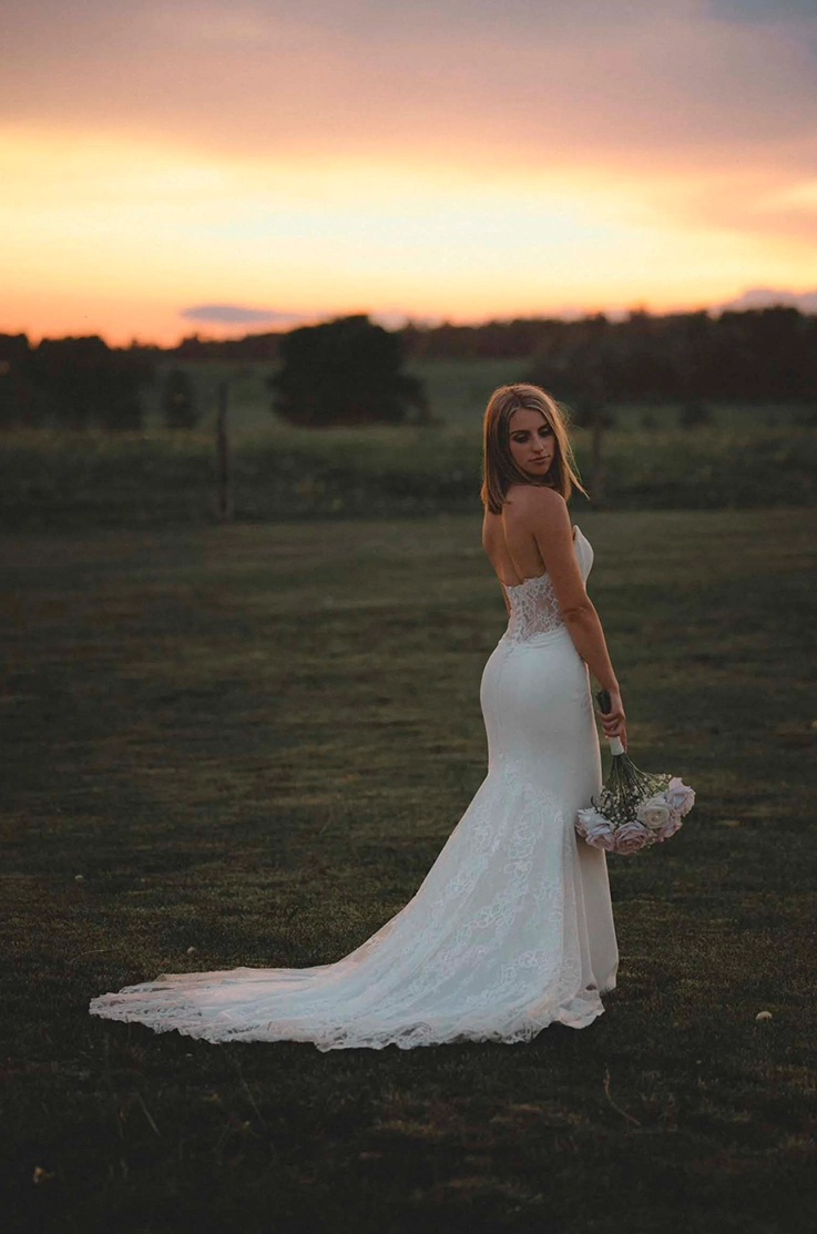 Bride poses in a strapless lace wedding dress with a crepe fit and flare skirt, holding a bouquet in an open field at sunset.