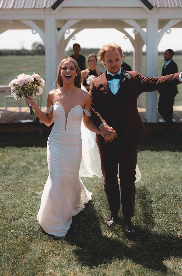Bride in a fit and flare crepe gown with a plunging neck, sleeves and veil, walks hand in hand with groom down the aisle.