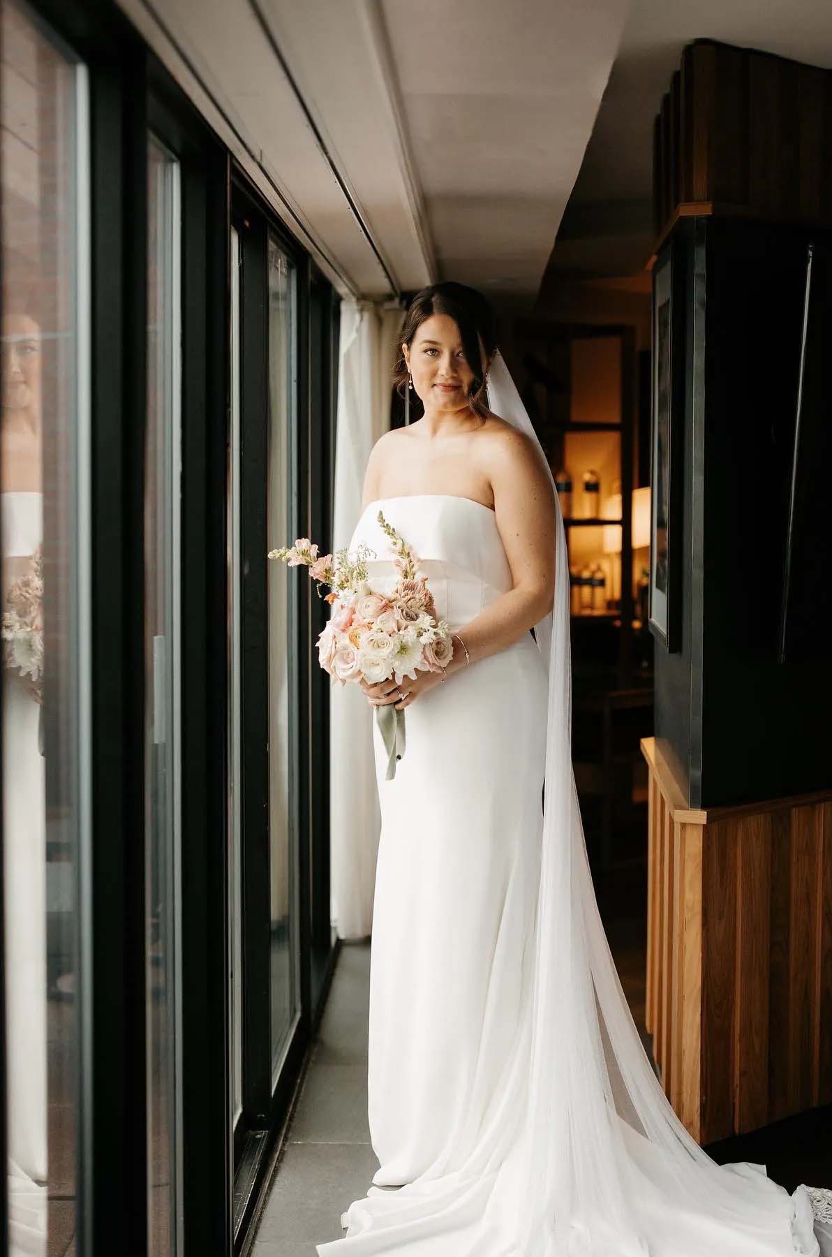  Bride poses in a hallway with black frame windows, in a crepe portrait collar gown with fit and flare skirt and a long veil.