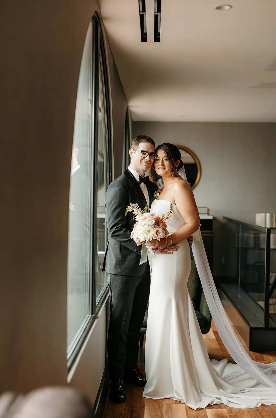 Groom embraces bride by windows, in a crepe fit and flare gown with a portrait collar and a veil, holding a blush bouquet.