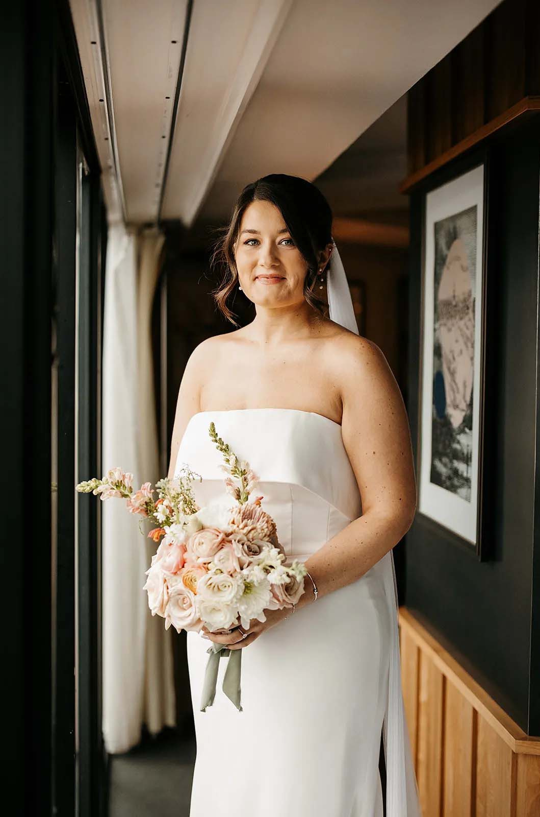 Close-up of bride posing in a hallway, in a crepe fitted gown with a portrait collar and a veil, and is holding a bouquet.