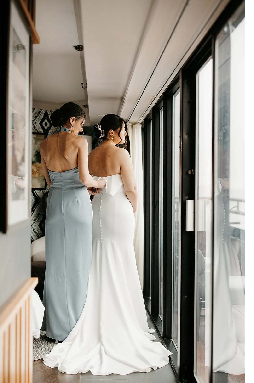Back of bride being fastened into a crepe fit and flare gown with buttons and a fold over neckline, by her bridesmaid.
