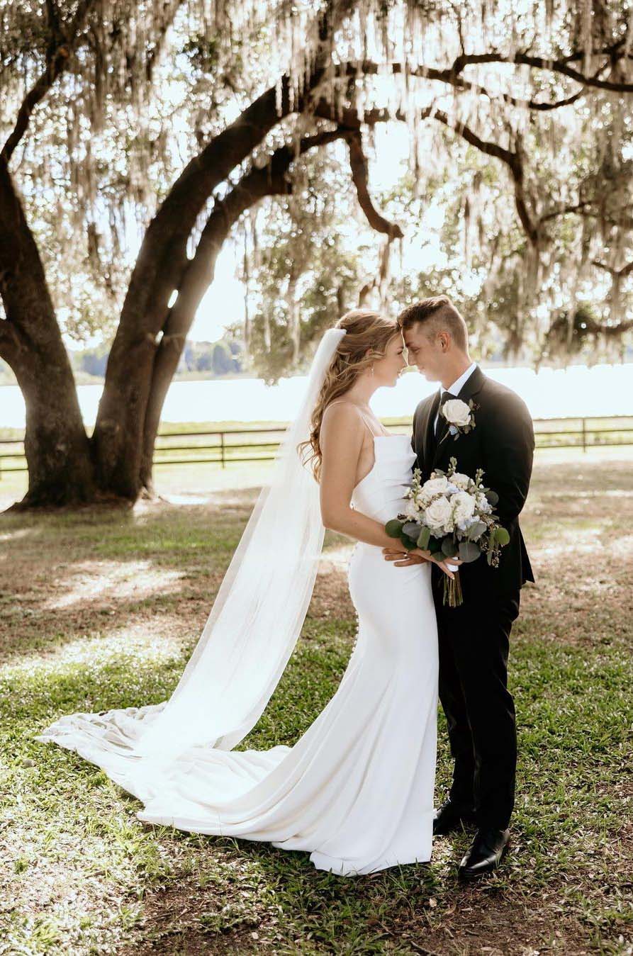 Groom embraces bride in a scoop neck crepe gown with buttoned fit and flare skirt and a veil, near a large willow tree.