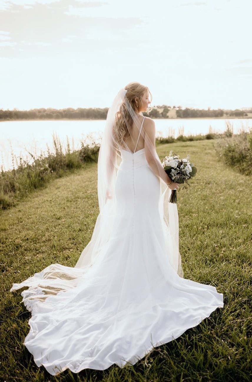 Bride poses in a crepe fit and flare wedding dress with a low back, thin straps and a veil, holding a bouquet at a lakeside. 