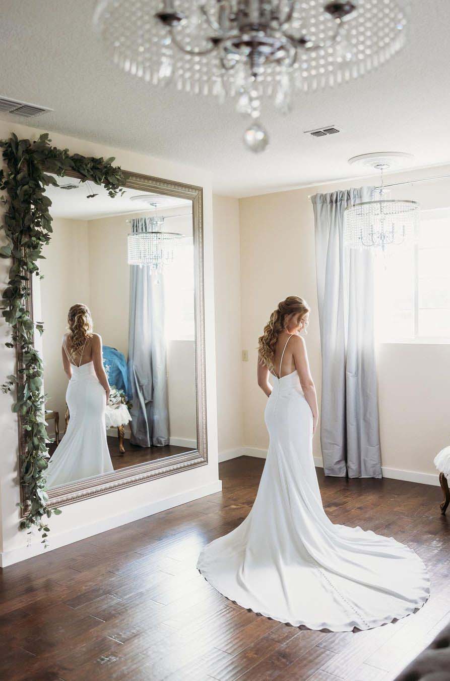 Bride in a fit and flare crepe gown with a strappy low back, stands in a bright room with a large mirror, under a chandelier.