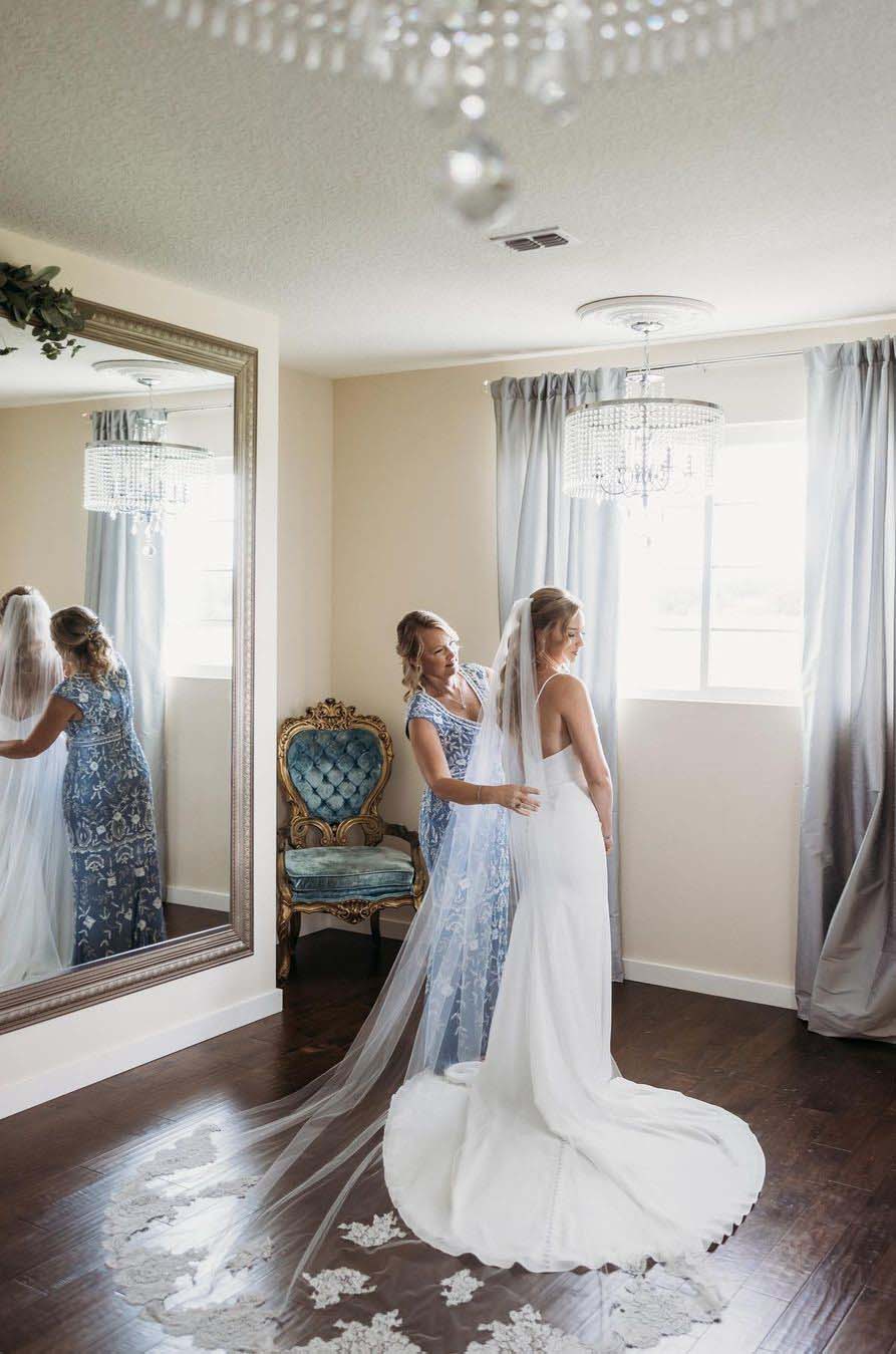 Bride gets ready in a brightly lit room, under a chandelier in a crepe fit and flare gown with a veil, as her mother helps