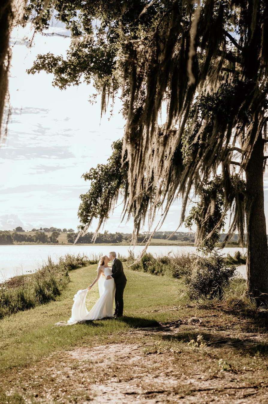  Groom kisses bride in a scoop neck fit and flare gown with a veil and she poses, under tall mossy oak trees by a lake.