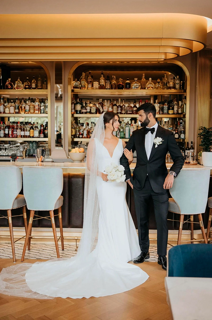 Bride in a plunging neck crepe gown with fit and flare skirt and a veil, poses beside her groom at a hotel bar.