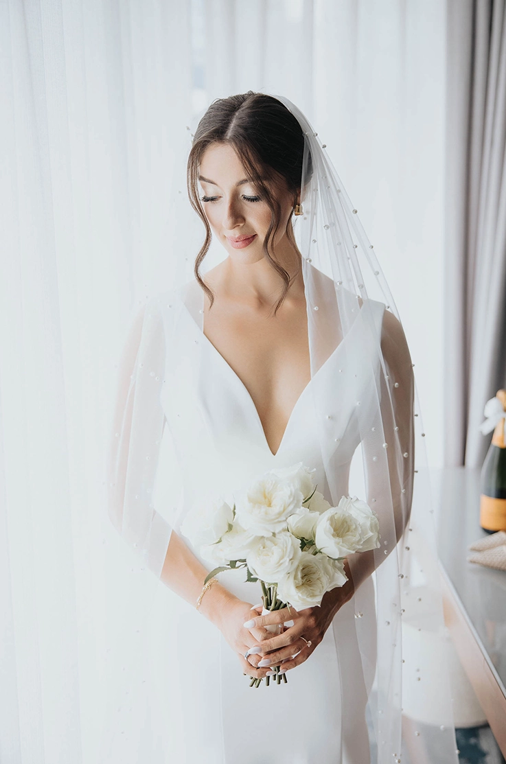 Close up of bride by a bright window, in a crepe fitted wedding dress with a plunging neck and a veil, holding a white bouquet.