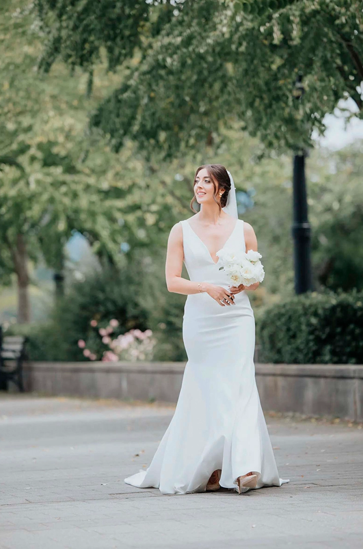 Brides walks on a tree-lined promenade in a crepe fit and flare gown with a plunging neck and veil, holding a bouquet.