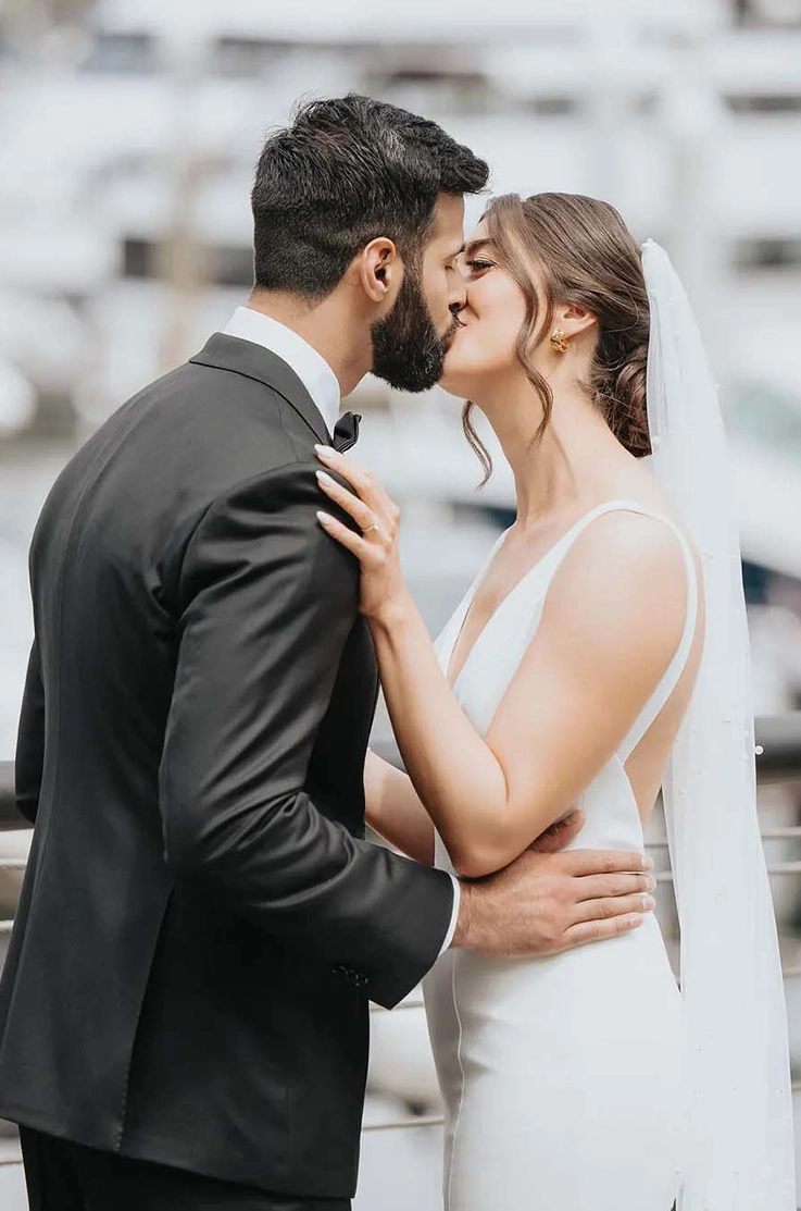 Groom kisses bride in a sleeveless crepe fitted gown with a plunging V-neck and a veil next to an outdoor railing.
