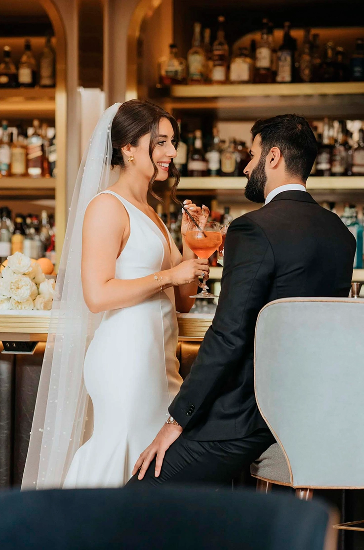 Bride in a deep-V neck crepe gown with princess seams and a pearl-edge veil, shares drinks with groom at a bar.