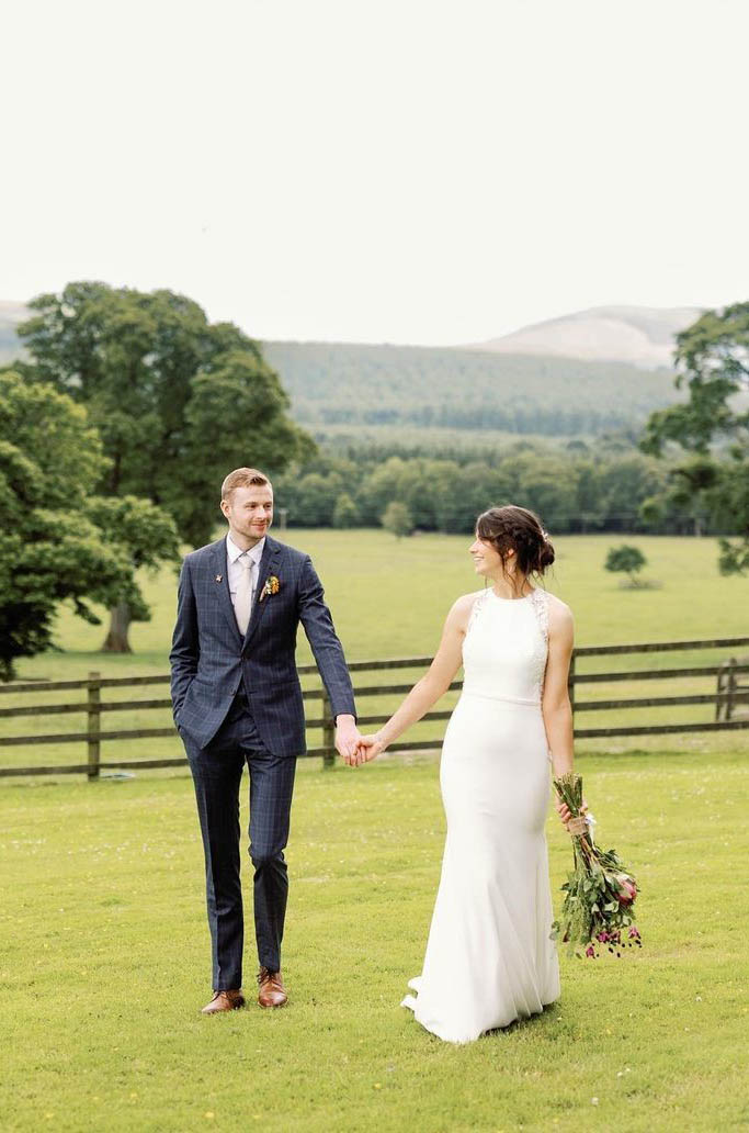 Bride in a sleeveless crepe and lace gown with a high neckline and belt, walks hand in hand with her groom in a countryside.